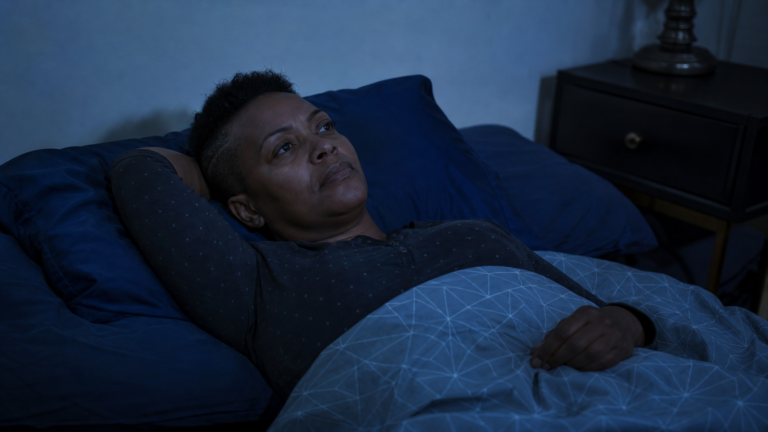 Mulher negra de 46 anos, cabelo curto raspado nas laterais, deitada na cama de olhos abertos olhando para o teto, expressão de cansaço sereno em quarto com luz azulada de madrugada.